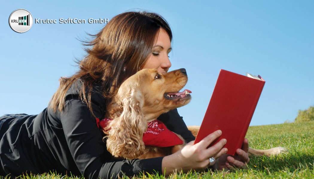 Eine junge Frau liegt bei strahlendem Sonnenschein mit ihrem Hund auf einer grünen Wiese und liest ein Buch